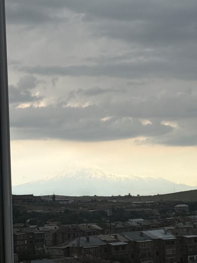 The image shows a view of Mount Ararat from Abovyan, Armenia. The foreground features residential buildings with dark roofs and multiple windows, indicative of an urban or suburban setting. Trees and other vegetation are visible between and behind the buildings. In the middle ground, there are more buildings and structures, including what appear to be industrial or commercial buildings on a hill. The background is dominated by the majestic snow-capped Mount Ararat, partially obscured by a hazy sky with layered clouds. The lighting suggests it is either early morning or late afternoon, with diffused light casting a soft glow. The overall scene depicts a landscape where human habitation coexists with a prominent natural landmark.