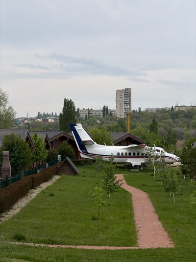 The image depicts a grounded, partially disassembled passenger plane resting on a green lawn among wooden structures, possibly cabins, in what appears to be a park or recreational area in the city of Kamyshin, Russia. A building under construction with a yellow crane is visible in the background. A gravel path runs alongside the airplane. The sky is overcast.