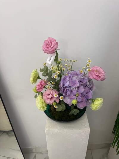 In Kaspiysk, Russia, a floral arrangement sits on a textured white pedestal. The arrangement features a large purple hydrangea as its centerpiece, surrounded by pink roses, pale yellow carnations, and delicate white daisies. A small tag labeled "Calin" is attached to one of the pink roses. The background is a plain white wall. To the left, a portion of a reflective surface, possibly a mirror or glass, is visible, showing a marble-tiled floor. To the right, a green fern plant is partially visible.