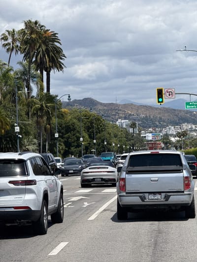The image captures a daytime scene on a multi-lane road in Beverly Hills, United States, under a cloudy, overcast sky. Traffic appears to be either stopped or moving slowly.
In the foreground, a white Jeep Grand Cherokee Laredo SUV is visible on the left, and a silver Honda Ridgeline pickup truck occupies the right lane. Between them, in the middle lane, a silver Porsche 911 convertible is prominent, with its top down. Further down the road, other vehicles, including a dark grey Tesla, are in traffic.
The road features clear white lane markings, including arrows indicating straight or left turns. Tall palm trees line the street on both sides, with dense green foliage creating a lush environment. Beyond the trees, rolling hills with sparser vegetation rise in the background. Several buildings are scattered across the hillsides, characteristic of the Southern California landscape.
Notable details include street lights, one of which holds a traffic signal displaying a green light. Adjacent to the traffic light, a "No U-turn" sign is clearly visible. A green street sign points left, with partial text reading "<- Palm Dr Beverly H," indicating "Palm Drive, Beverly Hills."
Visible text includes "LAREDO" on the white SUV. The license plate on the Porsche reads "DML 534" and on the Honda Ridgeline, "2O773U3." The word "HONDA" is also visible on the back of the pickup truck, along with "RIDGELINE." The combination of high-end vehicles, palm trees, and the visible street sign strongly places the scene in an affluent area like Beverly Hills.
