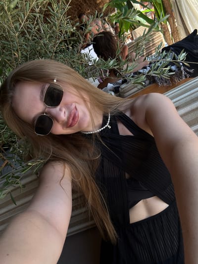 A young woman with long blonde hair and sunglasses takes a selfie. She is wearing a black dress and a pearl necklace. She is leaning against a tree with green leaves. In the background, there are other people and a striped cushion. The scene appears to be outdoors, possibly in a tropical or Mediterranean setting, and the presence of the striped cushion and the style of the foliage suggests a resort or beach location in Drobnići, Montenegro.
