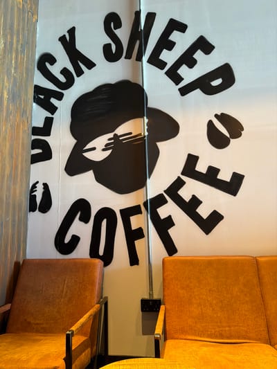 The image displays an interior view of a Black Sheep Coffee establishment located in Bretton, United Kingdom. A large black and white mural dominates the central white wall, featuring the text "BLACK SHEEP COFFEE" rendered in bold, hand-drawn style lettering. The "BLACK SHEEP" text arches above a stylized black silhouette of a sheep's head wearing sunglasses, with a coffee bean emblem positioned near its center and small coffee bean shapes flanking the text. A vertical silver metal conduit pipe runs centrally down the wall, partially obscuring the mural's sheep emblem, and terminates at a black double electrical socket mounted lower on the wall. To the left, a single armchair with orange-brown textured upholstery and a dark metal frame with wooden armrests is present. To the right, a matching two-seater sofa with similar upholstery and frame is visible. A segment of a light-toned, possibly wooden, round table appears in the lower foreground between the seating arrangements. A vertical strip of dark, weathered wooden paneling frames the left edge of the scene. No individuals are depicted.