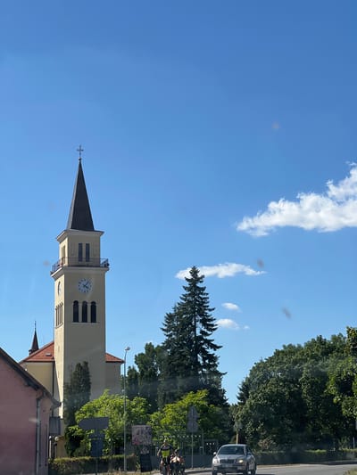 In Tvrdonice, Czechia, on a clear day with scattered clouds, a tall, light-yellow church tower with a dark conical roof and a cross at its apex dominates the scene. The tower features a prominent clock with Roman numerals, indicating a time between 5 and 6. Below the clock, arched openings reveal dark interiors, and a small balcony with railings encircles the clock face. To the left of the tower, a pink building with a red-tiled roof is partially visible. In the foreground, a gray sedan with license plate "3BE 4288" drives on a paved road. To the right of the road, several individuals are on bicycles, with two in the immediate foreground. Lush green trees and foliage surround the area, with a tall, dark green fir tree standing prominently near the church. A streetlamp is visible to the right of the church. The overall impression is a serene, daytime view of a European town.