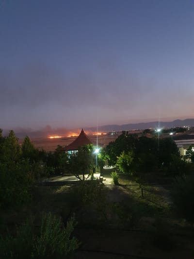 The image shows a dusk scene in Kheyrabad, Iran. In the foreground, there are several trees, some of which are illuminated by artificial lights. A pathway leads into the scene, with more trees and vegetation on either side. In the middle ground, there is a building with a conical roof, also illuminated by lights. Beyond the building, a large area is visible, with a distant fire or multiple fires burning, creating a hazy glow in the sky. In the background, there are mountains and a faint line of lights, suggesting a settlement or town. The sky is a gradient of deep blue and purple, with a few faint clouds.