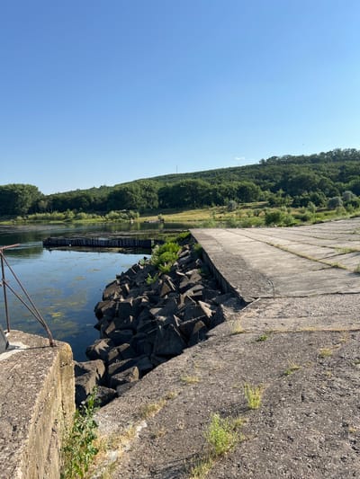 A concrete pier extends into a calm body of water, its surface cracked and weathered. The pier is lined on one side by a jumble of large, dark concrete blocks, forming a rough embankment. Beyond the embankment, the water is dotted with patches of green algae and a wooden structure, possibly a small jetty or barrier, is visible. On the far side of the water, a lush green hillside covered in trees rises gently under a clear blue sky. The scene is serene, with no people or animals immediately visible. The overall impression is one of a quiet, somewhat undeveloped waterfront.