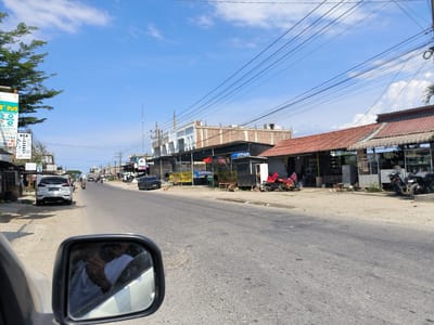 The image shows a street scene in Matang, Indonesia. The view is from inside a vehicle, looking forward. The street is asphalt with visible wear. Buildings line the right side of the street, including a two-story structure under construction and a roadside establishment with a red tile roof. A silver SUV is parked on the left side of the road. Other vehicles, including motorcycles, are visible further down the road. Power lines are strung overhead. The sky is blue with scattered clouds. The side mirror of the vehicle is visible in the foreground, reflecting a person. There is no indication of precise coordinates, time, or camera specifications.