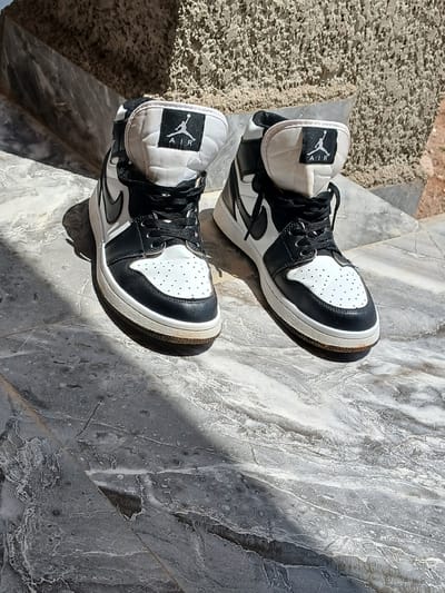 A pair of black and white Air Jordan 1 high-top sneakers are positioned on a marble-like surface. The sneakers are slightly dirty. The background consists of a rough, textured wall with brown and gray tones, and a diagonal shadow from an unseen object. The scene is illuminated by bright sunlight, creating distinct shadows on the surface. The location is Katon Rikkos, Nigeria.
