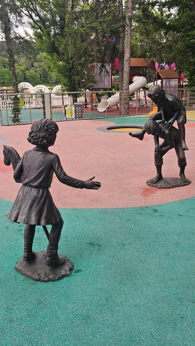 The image depicts a public playground located in Alikonovka, Russia. The playground features several bronze sculptures of children. In the foreground, a sculpture of a young girl, seen from the back, is holding a toy horse. Her right arm is extended forward, with her palm open. To the right, two other sculptures depict children engaged in play. One child is crouched down, and another is positioned on their back, appearing to be in a playful tussle. The ground of the playground is a soft, textured surface divided into two sections: a predominantly pink area and a turquoise-green area. In the background, elements of a playground structure with slides and colorful accents are visible, along with trees and foliage, suggesting a natural, park-like setting. A body of water and distant hills are visible beyond the playground.