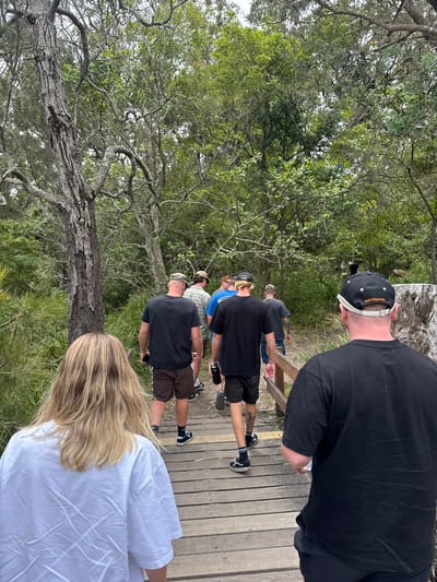 A group of people walk across a wooden walkway through a forested area. The walkway is elevated and surrounded by trees and lush greenery. Several men are in line, dressed casually in t-shirts and shorts, some wearing hats. A person with long blond hair, wearing a white shirt, is in the foreground. Another person, wearing a black shirt and a baseball cap, walks ahead. The scene appears to be a hike or walk in nature.