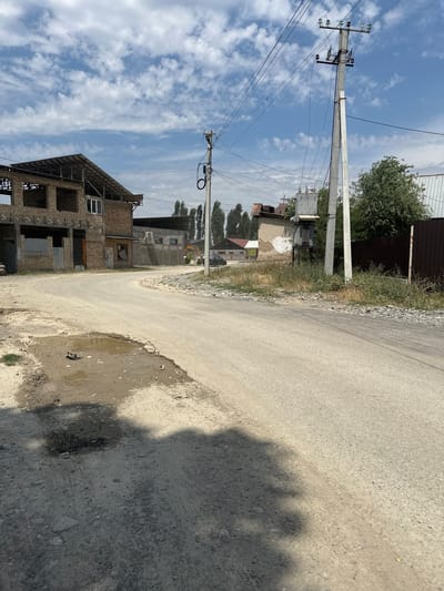 This is a vertical, medium-shot, outdoor photograph taken on a sunny day in Taygaraev, Kyrgyzstan. The scene depicts a rural street with a paved road curving to the right and a construction site on the left.
The road is rough and appears to have some water puddles on its surface. On the left side of the road, a multi-story brick building is under construction, with exposed rebar and unfinished window frames visible. Power lines and utility poles run along the street. Behind the construction site, several other buildings and trees are visible.
In the background, a large transformer sits near the utility poles. The sky is blue with scattered white clouds. The overall impression is one of a developing area on a bright, clear day. The photograph captures a sense of daily life in a semi-urban or rural environment. There is no visible text in the image.