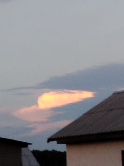 The image depicts a cloudy sky at dusk over a rural area in Крывасельскі сельскі Савет, Belarus. A prominent cloud formation in the sky is illuminated by the setting sun, casting a warm yellow and pink glow. In the foreground, the roofs of several houses are visible, one with a dark, corrugated metal roof and another partially obscured by shadow. The overall scene suggests a peaceful, twilight setting in a village.