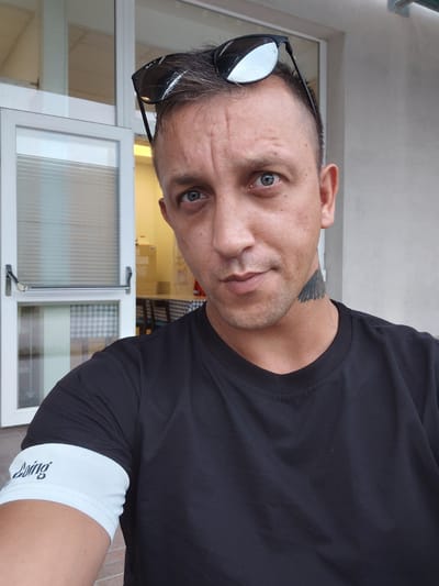 The image is a close-up selfie of a man with short, dark hair and blue eyes. He is wearing a black t-shirt and sunglasses perched on his head. A distinctive black tattoo of what appears to be feathers is visible on the right side of his neck. On his left arm, a white band with black lettering, possibly a brand name, is worn. The background suggests an outdoor setting, possibly near a building with a glass door and a wall. The lighting indicates it is daytime. The man is looking directly at the camera with a neutral expression. No specific location in Levico Terme, Italy, can be identified from the provided image.