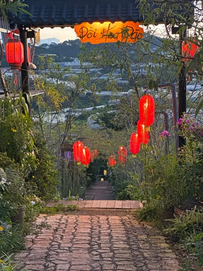 The image depicts a pathway lined with lush greenery and adorned with numerous red paper lanterns, creating a festive and inviting atmosphere. The pathway itself is paved with rough-hewn stones, suggesting an outdoor, perhaps rustic, setting. In the background, a valley unfolds with terraced landscapes, possibly rice paddies or tea plantations, hinting at a rural or mountainous region. The scene is illuminated by the warm glow of the lanterns, indicating that it is either dusk or evening. The lighting from the lanterns casts a soft, ambient light, and some are visibly lit from within.
To the left of the image, a traditional-looking roofline with dark tiles is visible, and an illuminated orange paper lantern with handwritten text hangs from it. The text is in an Asian script, likely Vietnamese given the location context of Lộc Quý, Vietnam. The overall mood is serene and tranquil, with a touch of cultural charm. The time of day is suggested by the soft light and the illuminated lanterns, likely sunset or early evening. The vegetation is abundant, with trees and bushes framing the pathway and lanterns. There are no people visible in the image. The setting appears to be a scenic outdoor space, possibly a garden, a restaurant, or a pathway leading to a scenic viewpoint.