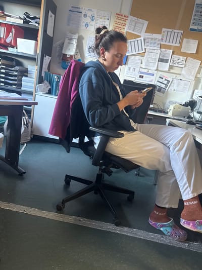 The image captures a person taking a break in an office or workstation setting, likely in Tourcoing, France. The individual, an adult with dark hair tied back, is seated in a black office chair, casually resting with their feet propped up while looking at a smartphone. They are dressed in a dark blue hoodie over a white top and white pants, possibly scrubs, which suggests a professional or medical work environment.
A notable detail is the person's footwear: brown socks with "JOLIE NANA" (French for "Pretty Girl") written on them, paired with colorful, tie-dye pattern Crocs-style shoes. The immediate surroundings include a desk with a computer monitor and other office equipment. The wall behind is covered with a corkboard and a whiteboard, both displaying numerous papers, charts, and documents. A prominent red sign on the corkboard contains French text, including "ALARME INCEN" (likely "ALARME INCENDIE" - Fire Alarm) and instructions such as "VERIFER LE NIVEAU CONCEN" (Verify the concentration level), "SUR LE REPORT D'ALARNE" (on the alarm report), "FIL S'AGIT DE SON MYGAU" (It's about its level), and "VERIFEZ DANS SON SERVICE" (Verify in your department/service). To the person's left, a shelving unit holds binders and files. The floor is a dark green or grey color. The overall scene depicts a relaxed moment in a functional office space.