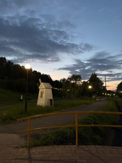 The image depicts a park scene in Vladikavkaz, Russia, during the twilight hours. A white, miniature windmill structure stands on a grassy slope, surrounded by dark trees. A pathway, likely a walking trail, curves through the park, bordered by lush green grass and a yellow railing. A streetlamp with a bright, glowing bulb illuminates the foreground, casting light on the path and nearby vegetation. The sky above is a dramatic mix of dark, brooding clouds and streaks of lighter hues, suggesting the transition from day to night. The overall mood is serene and peaceful, with a hint of atmospheric drama from the sky. There are no visible people or text in the image. The time of day is clearly evening, indicated by the low light and the artificial illumination.