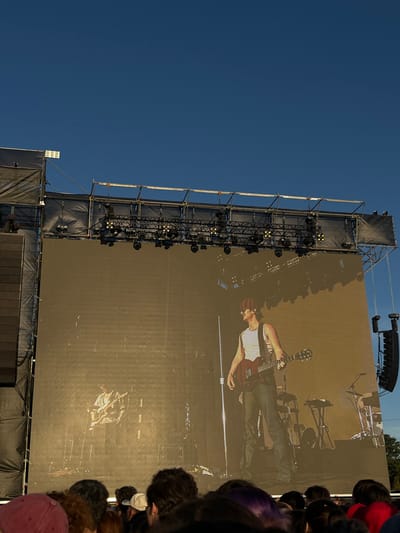 The image depicts an outdoor concert stage in San Isidro, Argentina, under a clear blue sky. A large, rectangular LED screen forms the central element of the stage, framed by a metal truss system above and on the sides. The truss supports numerous stage lights, some emitting a faint yellowish glow, and is partially clad in black fabric. Large, dark speaker cabinets are suspended from the right side of the stage structure, with a substantial, dark speaker array visible on the far left.
The LED screen displays a live video feed or pre-recorded image of a male musician. He is shown in profile, facing left, wearing a white tank top, dark pants, and a dark cap, while playing a dark red electric guitar. A second, less distinct male figure is visible to his left and lower on the screen, also appearing to play a guitar. Behind the primary musician on the screen, various music equipment, including keyboards on stands and elements of a drum kit, are visible.
The lower foreground of the image shows the backs of numerous heads belonging to the audience, indicating a crowd facing the stage. Hair colors vary, and some individuals wear caps, such as a reddish-pink one on the left. The scene is illuminated by natural daylight.