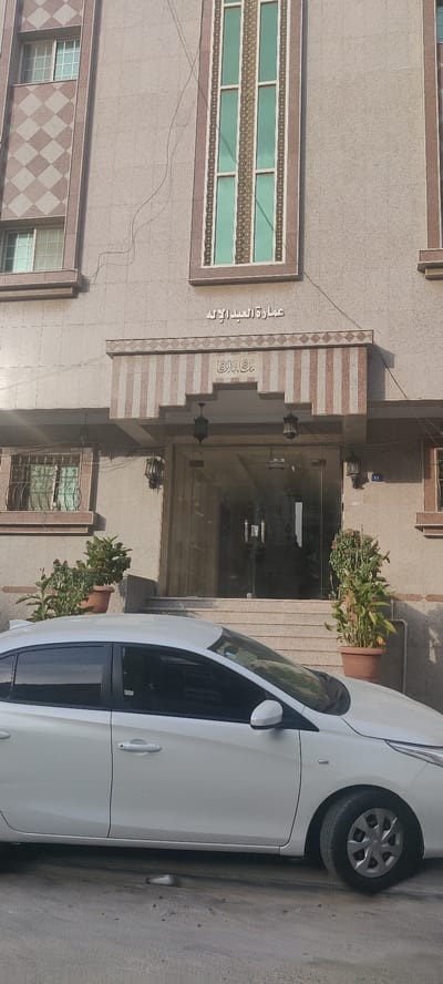 The image shows the exterior of a building, likely an apartment building, with Arabic text above the entrance, translating to "Emart Alabd Alilah". The building has a modern architectural style with a neutral color palette and decorative windows. In front of the building is a white sedan car parked on the street. There are potted plants flanking the entrance. The location is likely in Jeddah, Saudi Arabia, given the Arabic text and architectural style common in the region.