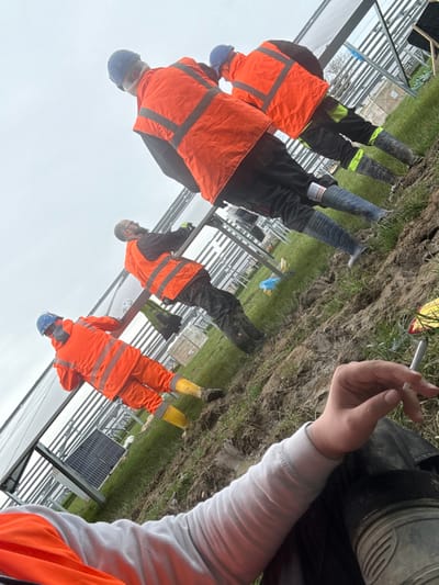 The image portrays an outdoor solar farm construction site in Baldurgen, Ireland, under an overcast sky. The ground consists of disturbed earth, mud, and patches of grass. Four individuals are present, all attired in personal protective equipment. This includes blue hard hats, orange high-visibility vests or jackets (one worker on the far left wears an orange high-visibility suit), dark work trousers, and rubber work boots (blue, black, and yellow).
Three individuals are positioned further from the camera, facing generally away, actively engaged with or standing near partially installed solar panel arrays mounted on metal racking structures. The worker furthest left appears to be handling or installing a solar panel.
In the immediate foreground, the right hand and forearm of a fourth individual are visible. This individual holds a lit cigarette between their fingers. A portion of their light grey long-sleeve shirt and an orange high-visibility vest are also visible.