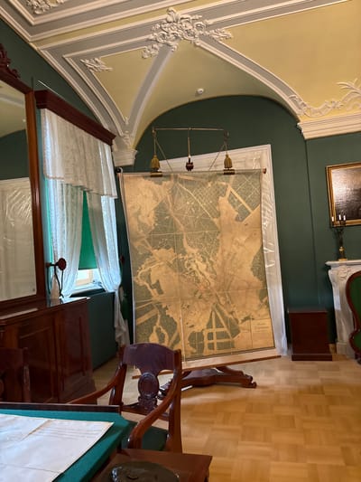 The image depicts an indoor scene, likely a museum or historical room, featuring a large antique map displayed on a wooden easel. The map is positioned in the center of the frame, leaning against a dark green wall. The map itself appears to be a detailed topographic or city plan, rendered in muted earth tones, with intricate lines and markings. The top of the map is secured by clips to a horizontal rod, which is suspended by wires and a decorative gilded finial.
To the left of the map, a tall, dark wooden cabinet with a mirror is visible. White lace curtains hang over a window behind this cabinet, and a green fabric is visible underneath. A dark wooden desk with papers on it is in the foreground, partially obscuring a dark wooden chair with an ornate backrest. To the right of the map, a white fireplace mantel is present, adorned with a framed painting above it and a candelabra. A small, dark wooden cabinet sits to the right of the fireplace. The room has a parquet wooden floor. The ceiling is arched and painted in shades of yellow and white, with ornate plasterwork.
The map has Russian text at the bottom, which reads "Планъ Гатчины" (Plan of Gatchina) and "съ обозначеніемъ улицъ и домовладеній" (with the designation of streets and properties). This indicates the location is Gatchina, Russia, and the map is a historical plan of the city. No people are visible in the image. The overall impression is one of a preserved historical interior, showcasing a significant cartographic artifact.