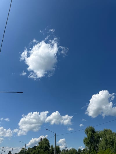This image captures a bright, clear blue sky with scattered white cumulus clouds. In the foreground, the tops of lush green trees are visible, indicating a natural or park-like setting. Several modern streetlights are positioned among the trees, suggesting an urban or suburban environment. A thin black electrical wire runs diagonally across the upper left portion of the frame, and another similar wire is visible in the upper right. The overall scene is tranquil and suggests a pleasant daytime view. The context provided places this scene in the city of Strășeni, Moldova.
