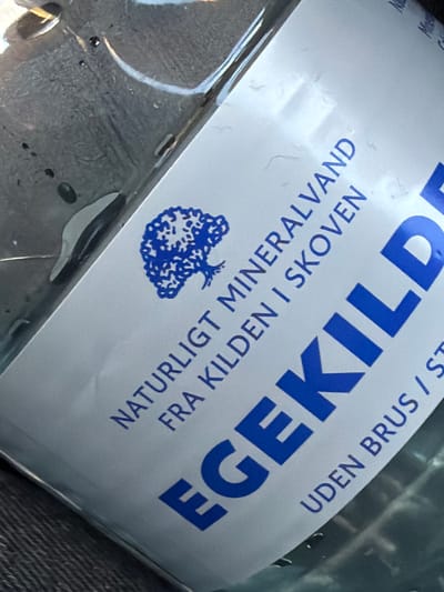 A close-up shot shows a label on a clear plastic bottle of mineral water. The label is predominantly white with blue text and a blue silhouette of a tree. The text on the label reads "NATURLIGT MINERALVAND FRA KILDEN I SKOVEN" and "EGEKILD / UDEN BRUS / ST". The bottle appears to be partially filled with water and is resting on a dark surface, possibly fabric or a car seat. The image is angled, with the label running diagonally across the frame. There are water droplets visible on the exterior of the bottle. The background is blurred, suggesting the photo was taken in a casual setting. There are no people or identifiable landmarks visible in the image. The city of Ödsmål, Sweden, is not depicted or referenced in the visual content of the image.