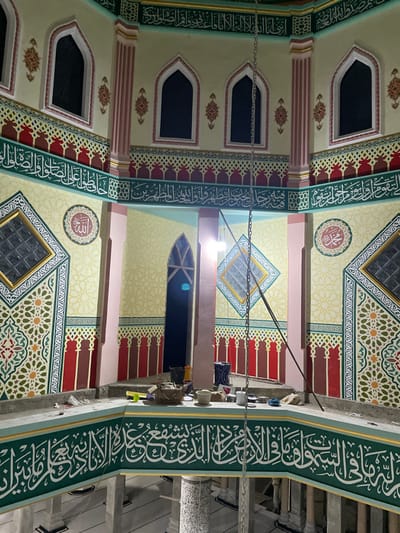This image displays the richly decorated interior of what appears to be a mosque in Cutneuheun, Indonesia, viewed upwards towards the ceiling and upper walls. The scene is illuminated by artificial lighting, indicating it is likely evening or night.
The walls and ceiling are adorned with elaborate Islamic art. Light cream-colored surfaces are bordered and segmented by prominent green bands featuring intricate white Arabic calligraphy. Interspersed throughout are complex geometric patterns in shades of red, brown, yellow, and green, creating a vibrant mosaic effect. One circular medallion with stylized Arabic script is also visible.
Several arched windows with dark panes are embedded in the walls, alongside square-shaped windows with textured glass integrated into the ceiling patterns. Metal chains traverse the space, likely intended for hanging light fixtures, although no specific lights are clearly attached at their ends. A narrow architectural ledge holds several small, decorative objects, possibly vases or cups. The overall impression is one of detailed craftsmanship and a serene, reverent atmosphere. No individuals are present in the image.