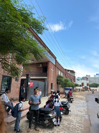 The image shows the exterior of "St. Agnes Mission Higher Secondary School" located in Jajmau, India. A group of people, including students in school uniforms and adults, are visible near the entrance of the school. Several students are carrying backpacks. Some individuals are standing, while others are seated on steps. A man in a blue shirt and dark pants is standing in the foreground, facing the camera. There are parked scooters and a three-wheeled vehicle on the paved area in front of the school. The building is constructed of red brick, and a white cross is mounted on the facade. The sky is blue with scattered clouds. The scene appears to be during the day.