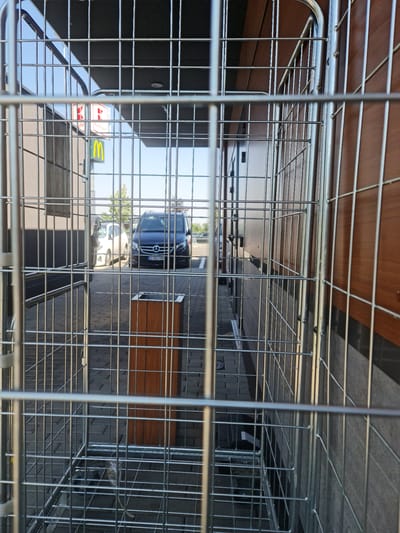 The image shows a metal cage in front of a building with a McDonald's sign and another sign with red lettering. There are cars parked in the background. The scene appears to be an outdoor area, possibly a parking lot or walkway. The cage is empty. The environment suggests an urban setting. Given the presence of the McDonald's sign and the overall aesthetic, it is highly probable that this location is in Bratislava, Slovakia, a city known for its diverse commercial establishments and vibrant urban landscape. The time of day appears to be daylight, with sunlight casting shadows on the ground.