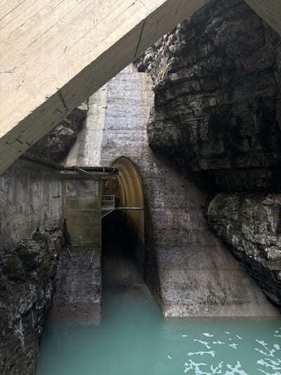 This image displays a concrete hydro-engineering structure situated within a natural rock gorge in Pellafol, France. A large, weathered concrete beam extends diagonally across the upper left portion of the frame. Below this, vertical concrete walls form a channel partially filled with still, turquoise-green water that occupies the lower half of the scene. On the right, a prominent arched concrete opening serves as the entrance to a dark, recessed tunnel. Inside this archway, an elevated metal walkway with handrails is visible, extending into the tunnel. The concrete surfaces exhibit extensive water staining, discoloration, and visible erosion. Natural, rugged rock formations with visible stratification flank both sides of the concrete structures, particularly prominent on the right. The overall environment is dimly lit, suggesting depth within the gorge. No individuals, animals, or active processes are visible.