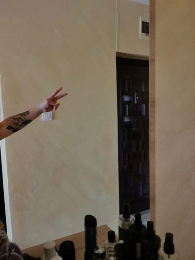 In a domestic interior, a person's tattooed arm, featuring a floral design, is partially visible on the left side of the frame. The hand extends towards the center, displaying a peace sign with bright yellow-painted fingernails. The arm rests against a beige-colored wall, to the right of which is a white light switch. The background reveals a dark, paneled door, framed by a light-colored partition. In the foreground, a collection of bottles, likely toiletries or cosmetics, is arranged on a wooden surface, with some bottles reflected in a mirror. The scene is situated in a room, implied to be a dwelling in the city of Aksakovo, Bulgaria.