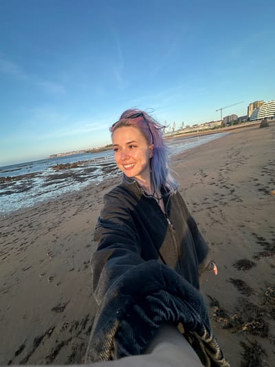 The image is a wide-angle selfie of a young woman with purple-tinted hair standing on a sandy beach in Gijón, Spain. She is wearing a dark denim jacket and a necklace. The beach has scattered seaweed and the tide is out, revealing rocky areas. In the background, the city of Gijón is visible with buildings and cranes, suggesting an industrial or port area. The sky is clear and blue with a hint of golden light from what appears to be sunrise or sunset. The woman is looking to her left and smiling. The perspective of the selfie places the viewer in a first-person viewpoint, as if standing next to her on the beach.