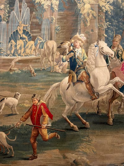 This is a detailed tapestry depicting a hunting scene in a garden setting. In the foreground, a young man in a red jacket and yellow breeches runs while holding a stick, leading two dogs. Behind him, a man on a white horse, wearing a hat and a blue jacket, holds a bird of prey. Another man on horseback, in green and brown attire, is partially visible in the background. The background features a fountain with a figure of Neptune, several other horses, and lush foliage, suggesting a park or palace grounds. The tapestry is likely displayed within the Royal Site of San Lorenzo de El Escorial, Spain, a historic royal palace and monastery.