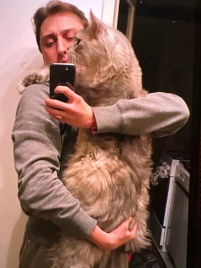A person is taking a selfie with a large, fluffy cat in what appears to be a residential bathroom. The person is wearing a gray sweatshirt and holding a black smartphone in their left hand. Their right arm is around the cat, which is held in a way that suggests it's being hugged. The cat is predominantly light brown and gray with very long fur. The background is partially visible, showing a light-colored wall on the left and a darker area on the right, possibly containing some household appliances. The image quality is somewhat blurry and grainy, as is typical of a low-resolution photograph taken in indoor lighting conditions. There is no visible indication of the location being in Verona, Italy.