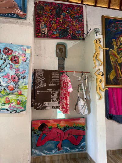 This image depicts an interior scene, possibly a shop or art studio, in Tejakula, Indonesia. The walls are textured white. On the left, a painting of a foot with the word "Nora" written on it is partially visible. Below it, a painting features pink and red circular shapes against a blue and green background, with a signature that appears to be "Syma".
Dominating the center of the image is a rack displaying clothing and bags. A pink and white patterned shirt hangs from a hanger, next to a white bag with a black and grey pattern. To the right of the rack, several brown fabrics are folded and hung, displaying text that includes "TWENTY BILLION DOLLARS" and "SOUTH AFRICAN RESERVE BANK".
On the wall above the rack, a colorful painting depicts figures with dark bodies and pink patterned backgrounds, possibly an abstract or narrative piece. To the right of this, a framed painting with dark colors and yellow and blue figures is visible.
Further to the right, two gold-colored decorative figures of human bodies are mounted on the wall. One is reaching upwards, while the other is in a dynamic pose.
Below the rack and to the right, a painting shows a red-skinned figure reclining in water, with a sunset sky in the background. The floor appears to be tiled. The ceiling is made of woven material, suggesting a tropical or traditional architectural style. The overall impression is of a space filled with art and textiles, possibly a commercial establishment.