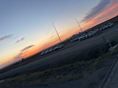 The image depicts a scene at dusk, likely at the Bozshakol mine in Kazakhstan. The sky is painted with hues of orange, pink, and blue, indicative of a sunset. Several white pickup trucks are parked in a row, some with decals that are illegible. They appear to be utility vehicles, possibly used for work at the mine.
In the background, a chain-link fence runs along a vast, open area. Beyond the fence, industrial structures and buildings are visible, their details softened by the fading light. Two tall lampposts stand out against the sky, one with a lit bulb. The ground is unpaved, a mix of dirt and gravel, with sparse vegetation growing in patches. A drainage grate is visible in the lower right corner of the image.
The overall atmosphere is quiet and serene, enhanced by the twilight setting. There are no people visible in the frame. The image captures a moment of transition between day and night, highlighting the industrial landscape against the natural beauty of the sunset. The location context of Bozshakol, Kazakhstan, suggests this is likely an operational area within or near the mining complex.