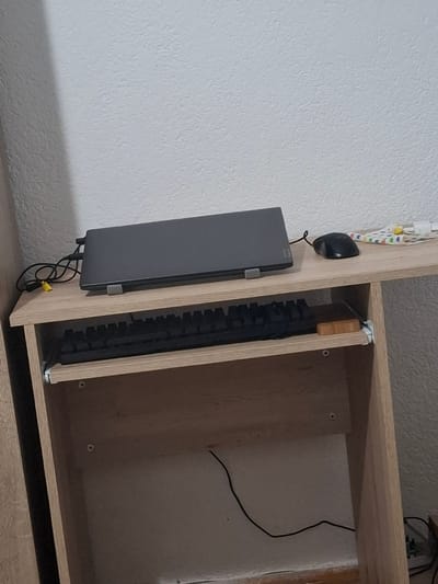 The image depicts a close-up view of a desk with a laptop, a keyboard, and a computer mouse. The desk appears to be made of light-colored wood. A gray laptop is closed and positioned on the right side of the desk. A black computer mouse is situated to the right of the laptop. Beneath the desk's surface, a black keyboard is visible. Cables from the laptop are seen dangling to the left. The background consists of a textured, off-white wall. The image does not contain any information about the city of Kran, Bulgaria, or any specific location within it. There are no people present in the image.