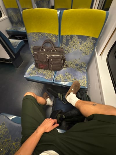 A person sits on a seat in a public transportation vehicle, likely a train. The seat has a yellow cushioned back and a blue seat with a repeating yellow flower pattern. A dark-colored briefcase sits on the seat to the left of the person. The person wears a brown clog with white socks and dark green shorts. A dark-colored backpack sits on the seat next to the person. The person's arm rests on their leg. The location is possibly Le Mesnil-Amelot, France, given the likely French origin of the train design and seating.