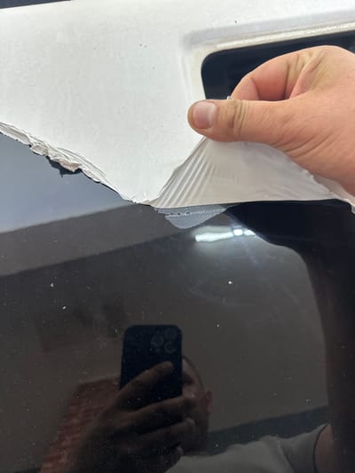 A gloved hand peels protective film from a white surface, revealing a black, possibly glossy surface beneath. The reflection of a phone in a hand is visible on the black surface. The location is unknown, and there is no evidence to indicate the scene is in Gabarevo, Bulgaria.