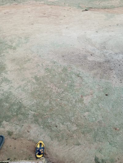 The image displays a dusty, weathered ground surface, likely a path or open area. The ground is primarily a light brown and grey color, with patches of faded green, possibly indicating moss or algae. Small stones and debris are scattered across the surface. In the lower left portion of the image, a black shoe is partially visible. To the right of the black shoe, a single yellow and blue sandal is lying on the ground. The sandal features cartoon characters and text that appears to read "Kiss" and "Cute". The overall environment suggests an outdoor setting in a dry or arid climate, with evidence of wear and tear on the ground. The scene appears to be located in Gindin Dutse, Nigeria.