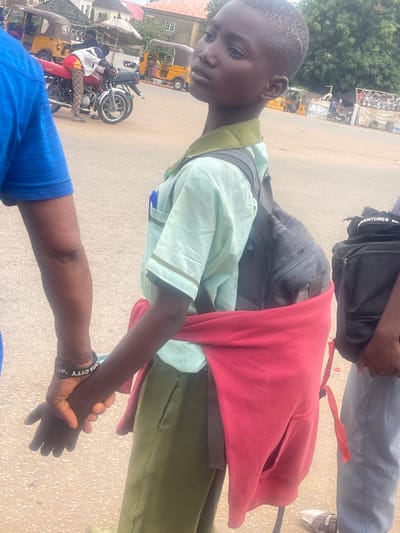 The image shows a young boy wearing a light blue shirt and a dark backpack. He is also wearing a red garment draped over his shoulders, and a green skirt or shorts. He is holding hands with another person whose arm is visible in the foreground, wearing a blue shirt. The boy appears to be looking to his left with a neutral expression. The scene is outdoors, with a dusty road and several vehicles in the background, including a motorcycle and two yellow auto-rickshaws. There is also some greenery and buildings visible. The overall lighting suggests it is daytime. A black backpack with the word "ADVENTURES" is visible at the top of the image. Another person's legs are also visible, wearing grey trousers and white sneakers. A black wristband on the hand holding the boy's hand has "OFA CITY W" written on it. The context provided indicates the location as Galadimawa Village, Nigeria.