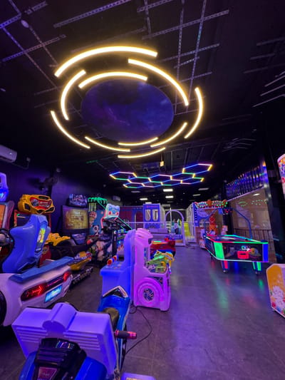 The image depicts an indoor arcade or entertainment center, characterized by its dark ambiance illuminated by vibrant neon and LED lights. The scene is dominated by various arcade games arranged in rows. In the foreground and extending to the right, several shooting games are visible, featuring colorful housings in blue, white, and purple, equipped with light-up gun controllers. Further back, other game machines, possibly including racing games, can be seen with their glowing screens and interactive setups.
The left side of the frame showcases prominent decorative lighting. A large, dark, circular object, possibly representing a planet or portal, is encircled by bright, glowing yellow curved light strips that create a dynamic visual effect. Above and to the right of this, a pattern of interconnected hexagonal light fixtures glows in various colors like blue, green, and purple, contributing to a futuristic aesthetic. The floor is a dark, hard surface, reflecting some of the colorful lights from the games and wall installations. The ceiling is dimly lit, revealing some structural elements.
There are no people visible in the image, implying the area might be empty or photographed during off-peak hours. The arrangement of numerous game machines clearly indicates a setting for gaming and entertainment activities. The overall atmosphere is created by the low light conditions and the strong contrast of the colorful, glowing elements.
Visible text includes "ULTRA FIREPOWER" prominently displayed on one of the game screens, and "HIGH SCORE" on another screen, along with other unreadable game-related text. Although the context provided is Chennai, India, there are no specific visual cues within the image that uniquely identify this location.