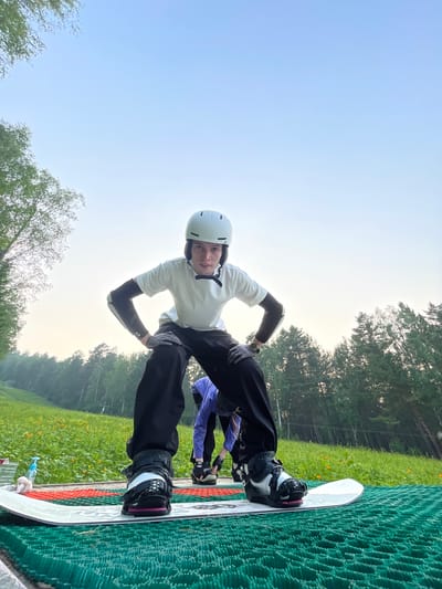 A low-angle, full shot captures a young person in a snowboarding stance on a green mat, with a snowboard beneath their feet. The person is wearing a white helmet, a white t-shirt, black pants, and grey gloves, with elbow pads visible on their arms. Their stance is wide, with knees bent and hands on their hips. In the background, a grassy slope dotted with yellow flowers leads up to a dense forest. The sky is clear and pale blue, with the soft light suggesting early morning or late evening. The location is the Divnogorsk Ski Resort in Divnogorsk, Russia, a place known for its natural beauty and outdoor recreational activities.