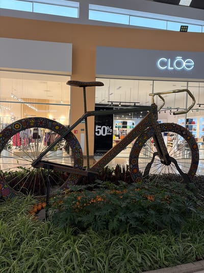 The image depicts a large, ornamental bicycle sculpture in front of a "CLOE FACTORY STORE" at an indoor shopping mall location in San Francisco Ocotlán (Ocotlán), Mexico. The bicycle is intricately decorated with colorful, possibly Huichol-inspired patterns. The bicycle sculpture is surrounded by dense greenery. Inside the store, a "50% off" promotional sign is visible.