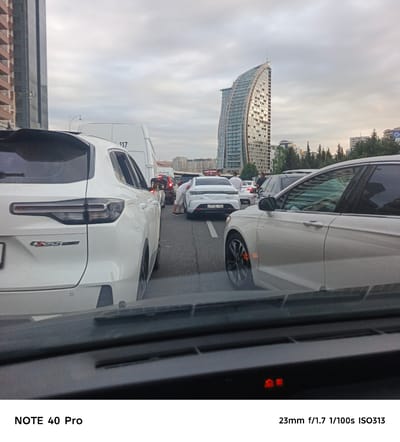 The image captures a traffic jam on a street in Khatay Raion, Azerbaijan, with several cars visible. A white SUV is in the foreground, to the left, with its rear facing the camera. To its right, a white sedan is stopped. Behind these, other vehicles are seen, including a white bus with the number "117" on its side. In the background, modern buildings, including a distinctive curved skyscraper, stand against an overcast sky. The scene appears to be a typical urban environment with a high density of vehicles.