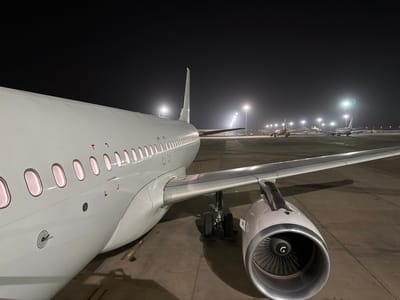 At night in what appears to be Blaise Diagne International Airport in the city of Communauté rurale de Diass, Senegal, an airplane, likely an Airbus A320 family aircraft, is parked on the tarmac. It is white, with visible windows, a wing, and one engine. Other aircraft are visible further in the distance under bright runway lights. The ground is concrete. The sky is dark.