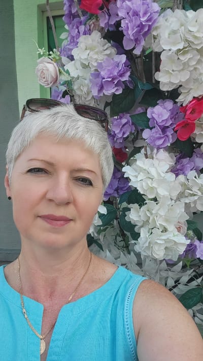 In Belaazyorsk, Belarus, a woman with short, silver hair and sunglasses perched on her head poses for a selfie against a backdrop of artificial flowers. She is wearing a turquoise shirt with intricate trim on the shoulders. The flowers are a vibrant mix of white, purple, and pink hydrangeas, with a few pink roses interspersed. The background includes a pale green building wall and a hint of a window. The woman has a light complexion and dark eyeliner. She is wearing a gold necklace with a heart-shaped pendant. The lighting suggests an outdoor daytime setting.