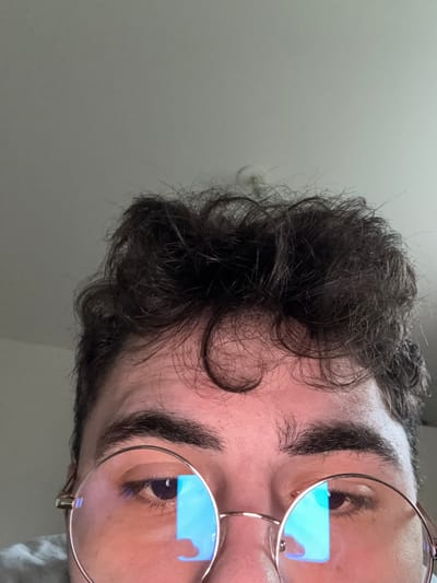 The scene is located in Rennes, France. The image provides a close-up, low-angle view of a person's upper face. The subject has dark, curly hair styled above the forehead and prominent dark eyebrows. The individual is wearing round, gold-rimmed eyeglasses. The lenses of the eyeglasses exhibit distinct blue, rectangular reflections, consistent with light emitted from a digital screen. The subject's eyes are partially visible behind the lenses. The background features a plain, light-colored ceiling. A circular, white ceiling fixture or detector is partially visible above and behind the subject's hair, in the upper right quadrant of the image. A minimal portion of light-colored fabric, likely clothing, is visible at the extreme bottom left.