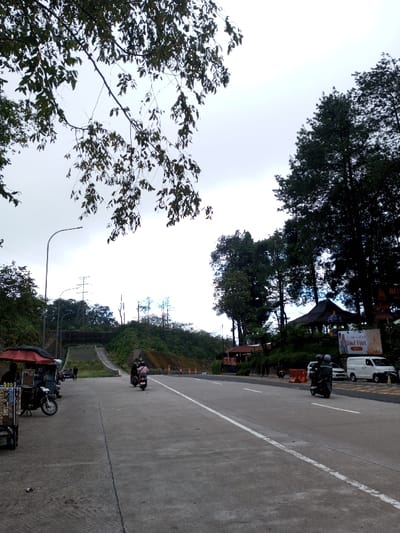The image depicts a paved two-lane road in Phoxay, Laos, under an overcast sky. In the foreground, a branch with dark green leaves extends from the upper left. The road, marked with a solid white line on the left shoulder and a broken white line dividing lanes, recedes into the distance.

On the left side of the road, a vendor cart with a red umbrella canopy is positioned on the shoulder. Merchandise is visible on shelves within the cart. A dark-colored motorcycle is parked next to the cart, facing away from the viewer. Further down the road, a streetlight pole curves over the road. Beyond this, a hillside with dense green foliage has been cut, revealing an embankment and utility poles in the background. A light-colored motorcycle with a single rider is visible on the road in this area, moving away from the camera.

On the right side of the road, tall evergreen trees flank a traditional-style building with a dark, multi-tiered roof. Below the building, a large banner displays text, including "Idul Fitri". Orange traffic barriers are placed along the road's edge. A black motorcycle with two helmeted riders is prominent on the road, traveling away from the camera. Behind the barriers, a white van and a partially visible white car are parked. A dark object, possibly refuse, is on the ground near the left shoulder in the immediate foreground.
