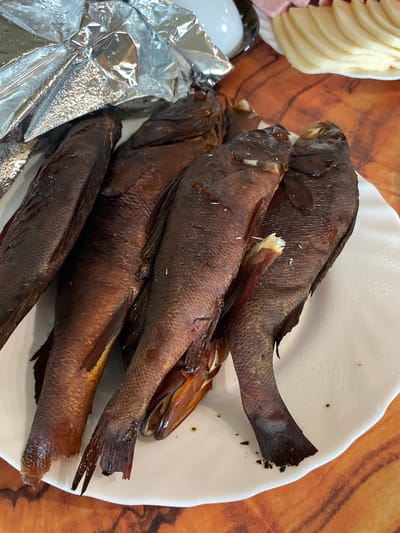 The image shows a plate of smoked fish. There are at least four fish on the plate, arranged head-to-tail. The fish are dark brown and appear to be whole, with scales visible. Some of the fish have their mouths open, revealing their insides. A piece of aluminum foil is draped over the top left of the plate, partially covering the fish. The plate is white and has a slightly raised edge. The plate is set on a wooden table with a reddish-brown, wood grain pattern. In the background, to the right of the plate, there is a platter with sliced cheese and what appears to be some pink meat, possibly ham. A white bowl is also visible in the upper left corner. The lighting is bright and suggests an indoor setting. The image is a close-up shot, focusing primarily on the food. There are no people visible in the image. The scene is consistent with a meal being served or prepared. The location is not specified in the image, so its inclusion in Solov'evka, Russia is external information not derived from the visual data.