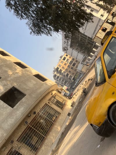 A tilted perspective captures an urban scene, dominated by a yellow vehicle in the upper portion. Buildings in various states of construction line a street, some with scaffolding and incomplete facades. A tree with dense foliage is visible on the left. The lower portion of the image shows a building with exposed window openings and a facade with barred windows. The overall composition evokes a sense of urban development and everyday city life.